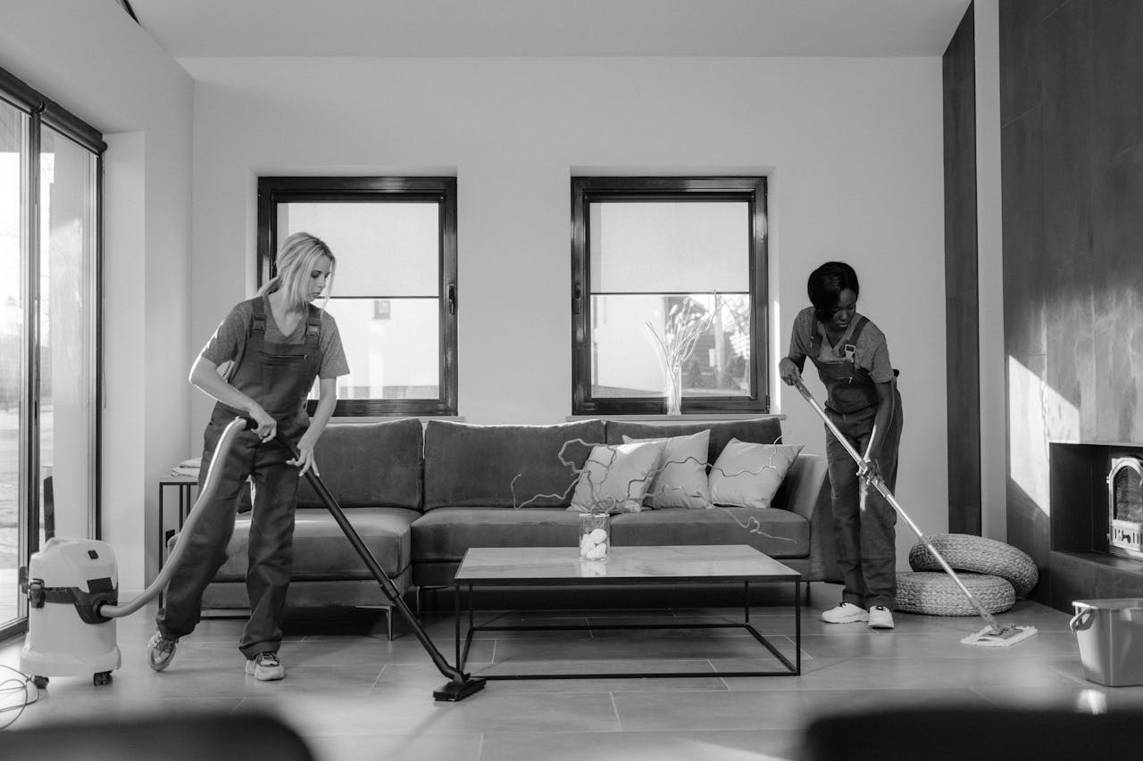 services-02 Two women in work clothes cleaning a modern living room using a vacuum and mop.